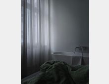 Ein minimalistisch eingerichtetes Schlafzimmer mit einer freistehenden weißen Badewanne neben einem großen Fenster mit halbtransparenten Vorhängen; im Vordergrund liegt ein Bett mit grüner Bettdecke.