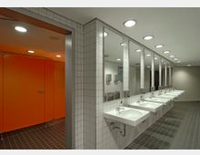 Öffentlicher Waschraum mit weiß gefliesten Wänden und Boden aus dunklen Fliesen. Auf der rechten Seite befinden sich mehrere Waschbecken mit Spiegeln darüber, links sind leuchtend orangefarbene Toilettenkabinentüren zu sehen.