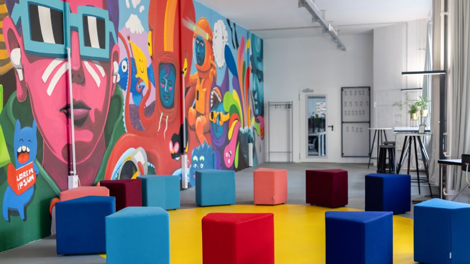 Großer, moderner Veranstaltungs- oder Gemeinschaftsraum mit hoher Decke und sichtbaren Schienen, an einer langen Wand mit farbenfrohem Graffiti-Mural. In der Mitte steht ein Kreis aus bunten Würfelhockern auf einem gelben Rundfeld; am Rand befinden sich Stehtische, Vorhänge und große Fenster.