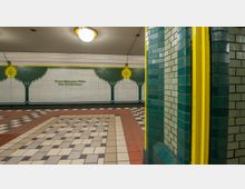 U-Bahn-Station mit dekorativen Wandfliesen in Grün- und Gelbtönen, die Bäume und gelbe Ornamente darstellen, sowie geometrisch gemustertem Bodenbelag in Rot- und Beigetönen. Im Vordergrund ist eine grün-gelbe Fliesenmauer zu sehen.