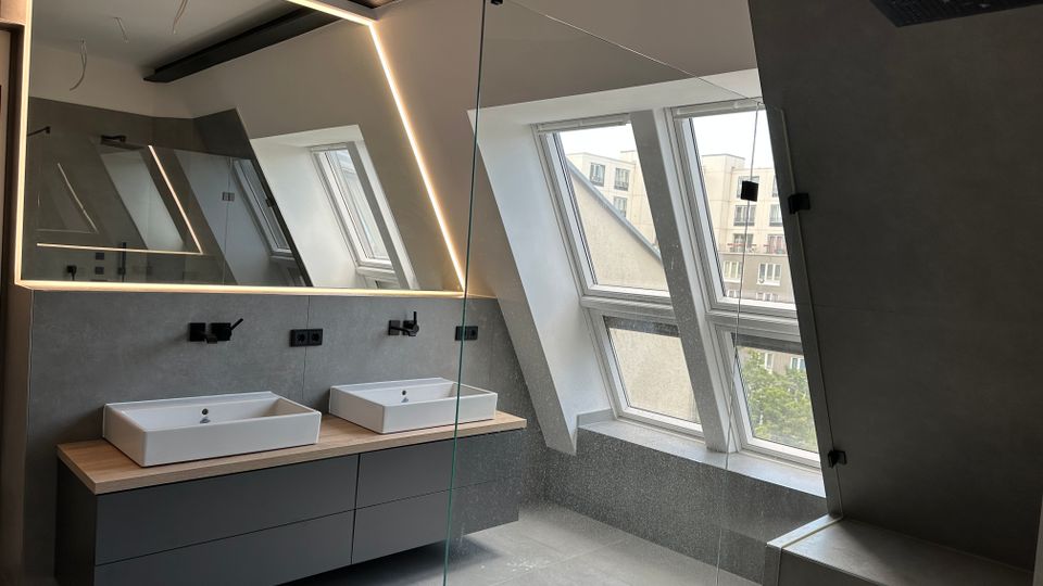 Modernes Badezimmer mit schräger Dachdecke, großem LED-beleuchtetem Spiegel und Doppelwaschtisch auf einem grauen Unterschrank. Rechts eine verglaste Dusche und mehrere Dachfenster, die Tageslicht und Blick auf umliegende Gebäude bieten.