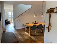 Esszimmer in einem modernen Zuhause mit einem Holzboden, einem Esstisch aus Holz und grauen Stühlen. Im Hintergrund führt eine Treppe nach oben, links ein Bereich mit großen Fenstern und Blick auf den Garten.