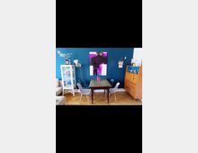 Wohnzimmer mit einer dunkelblauen Wand, auf der ein großformatiges abstraktes Kunstwerk in Lila- und Schwarz-Tönen hängt. Im Vordergrund steht ein Holztisch mit vier modernen Stühlen, daneben befinden sich eine Glasvitrine links und eine hölzerne Kommode rechts. Der Raum hat Holzboden und dekorative Details wie Kerzen und Pflanzen.