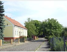 Gelbes Einfamilienhaus mit rotem Ziegeldach, umgeben von einem Zaun und einem kleinen Vorgarten, steht an einer gepflasterten Straße, die von Bäumen und einem Maschendrahtzaun gesäumt ist. Im Hintergrund ist dichte Vegetation sichtbar.