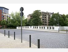 Straßenecke mit gepflastertem Gehweg und Asphaltstraße, im Hintergrund ein mehrstöckiges Wohngebäude, eine Baumreihe und eine weiße Brücke mit Gittergeländern. Im Vordergrund stehen schwarze Metallpoller und ein Verkehrsschild auf einem Mast.