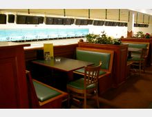 Ein Innenraum einer Bowlingbahn mit Sitzbereichen aus Holz und Polsterbänken in grünem Kunstleder. Im Hintergrund sind mehrere Bowlingbahnen und darüber an der Decke hängende Bildschirme zu sehen, dekoriert mit hängendem Weihnachtsschmuck.