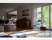 Wohnzimmer mit einem hellen Holzboden und großer Fensterfront, durch die ein Garten zu sehen ist. Im Raum befinden sich ein Holzschrank, ein Bücherregal, ein Esstisch mit Stühlen und eine Hängelampe; Tageslicht fällt in den Raum.