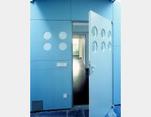 Tür eines modernen Gebäudes mit hellblauer Fassade, verziert mit runden Fensterausschnitten. Die geöffnete Tür gibt den Blick auf einen schmalen Flur mit dunklem Fußboden und einem hell beleuchteten Raum im Hintergrund frei.