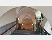 Innenansicht eines Treppenhauses mit breiter Steintreppe, dunklen Granitsäulen und einem hohen, weiß verputzten Kreuzgewölbe. Runde Wandlampen und ein grünes Geländer rahmen den Aufgang zu einem gewölbten Flur.