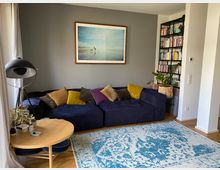 Wohnzimmer mit blauem Sofa, das mit bunten Kissen dekoriert ist, vor einer grauen Wand mit gerahmtem Bild. Ein runder Holztisch mit Dekoobjekten und eine Stehlampe stehen links, rechts befindet sich ein schwarzes Bücherregal mit Büchern und Pflanzen. Ein blauer Teppich bedeckt den Holzboden.