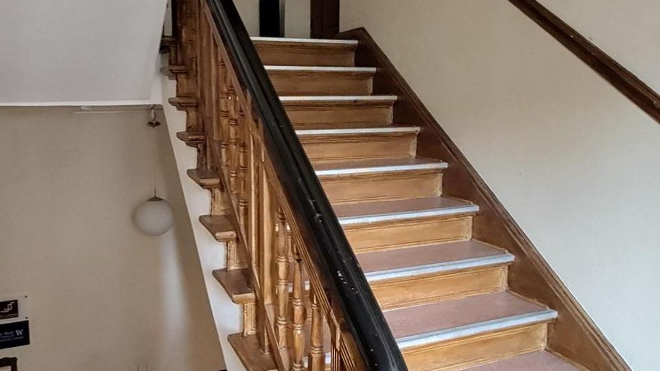 Treppenhaus in einem älteren Gebäude mit Holzstufen und gedrechseltem Holzgeländer, dessen Handlauf schwarz lackiert ist. Helle Wände, ein Zwischenpodest und eine hohe Tür am oberen Absatz prägen den schmalen, mehrstöckigen Innenraum.