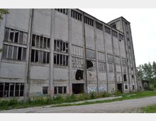 Verlassenes, mehrstöckiges Industriegelände aus grauem Beton mit zahlreichen kaputten oder fehlenden Fensterscheiben. Die Fassade ist teilweise mit Graffiti bedeckt, und um das Gebäude herum wächst überwuchertes Gras.