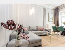 Helles, modernes Wohnzimmer mit großem grauen Ecksofa, Zierleisten an der Wand und einem gläsernen Couchtisch auf hellem Holzboden. Rechts schließt ein Essbereich mit grünen Stühlen an, neben bodentiefen Vorhängen und einem großen Fenster.