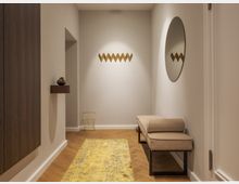 Minimalistisch eingerichteter Flur mit hellen Wänden, Holzboden und einem gelb gemusterten Teppich. Auf der rechten Seite befinden sich eine beige Sitzbank und ein runder Spiegel, während links ein schwebendes Wandregal mit einer dekorativen Figur zu sehen ist.