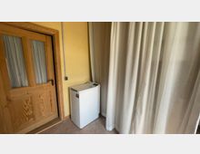 Ein kleiner Raum mit einer Holztür mit Glaseinsätzen auf der linken Seite, einer gelben Wand und einem weißen Minikühlschrank in der Ecke. Rechts hängen helle Vorhänge, die einen Teil des Raumes abdecken.