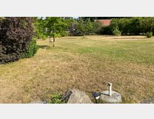 Weitläufige Wiese mit vereinzelten Bäumen, Sträuchern und einem freistehenden Gartenobjekt; im Hintergrund sind Häuser mit roten Dächern und Hecken zu sehen. Im Vordergrund liegen Felsen und eine kleine Metallleuchte auf einem Betonsockel.