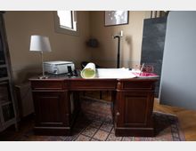 Ein traditionelles Arbeitszimmer mit einem dunklen Holzschreibtisch, auf dem eine Tischlampe, ein Drucker, ein Kartendokument und ein Modell eines Flugzeugs stehen. Im Hintergrund befinden sich ein Fenster, ein grauer Raumteiler und ein eingerahmtes Weltkartenbild an der Wand.