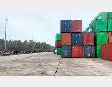 Ein Lagerplatz mit gestapelten roten, grünen und blauen Frachtcontainern auf einer Betonfläche, daneben Schienen mit leeren Güterwaggons und im Hintergrund eine Baumreihe unter bewölktem Himmel.