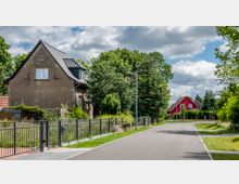 Wohnstraße mit Einfamilienhäusern und Vorgärten, darunter ein leicht verwittertes Gebäude links mit Schrägdach und ein rotes Haus im Hintergrund. Die Straße ist asphaltiert, von Gehwegen und Zaunanlagen gesäumt, und von Bäumen und Sträuchern umgeben.