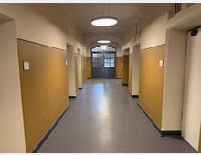 Ein langer, geradliniger Flur in einem Gebäude mit gelben Wandpaneelen und cremefarbenen Wänden. Türen führen zu beiden Seiten, und am Ende des Flurs befindet sich eine große Glastür mit Metallrahmen unter einem gewölbten Fenster.