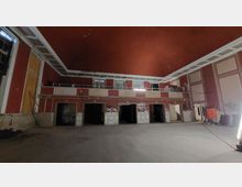 Großer, leerer Innenraum eines verfallenen Gebäudes mit roten Wänden und cremefarbenen Akzenten, hoher Decke und einer Empore mit Geländer. Im unteren Bereich befinden sich mehrere Türöffnungen, teils mit Graffiti bedeckt, und der Boden ist unbebaut.