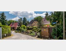 Wohngebiet mit einer schmalen Straße, gesäumt von Einfamilienhäusern mit roten und braunen Dächern. Vorgärten mit dichten Grünpflanzen, Zäunen und einem Straßenschild mit der Aufschrift „Furcher Str.“.