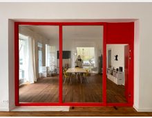 Moderne Innenraumaufnahme eines Büros oder Besprechungsraums mit einer roten Glastrennwand. Der Raum hat Holzboden, weiße Wände, große Fenster mit Vorhängen, ein Besprechungstisch mit gelben Stühlen und Regale an der Rückwand.