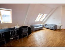 Heller Raum mit Dachschrägen, Holzboden und mehreren Dachfenstern, ausgestattet mit einem langen Tisch mit schwarzer Tischdecke, klaren Kunststoffstühlen, grauen Sofas und Blumendekoration.