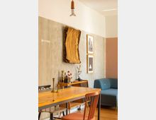 Wohnbereich mit modernem Einrichtungsmix: Ein Holztisch mit Stühlen im Vordergrund, eine blaue Sofaecke rechts, eine Wand mit goldener Kunstinstallation und gerahmten Grafiken sowie ein Regal mit Flaschen und Dekoration im Hintergrund. Der Raum hat eine minimalistische Beleuchtung und einen Holzboden.