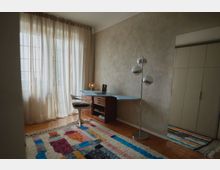 Ein minimalistisches Arbeitszimmer mit einem geschwungenen Schreibtisch in Holz und Blau, einem modernen weißen Stuhl und einer silbernen Stehlampe mit kugelförmigen Leuchten. Ein großer Kleiderschrank mit hellen Türen steht rechts, der Raum ist durch bodenlange, durchscheinende Vorhänge und einen bunten Teppich akzentuiert.