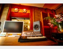 Ein Musikstudio mit einem Synthesizer auf einem Holztisch, einem Lautsprecher, einem Computerbildschirm sowie roten Schallabsorbern an der Wand. Im Hintergrund ist ein Fenster mit Blick in einen anderen Raum und eine Vase mit Blumen sichtbar.