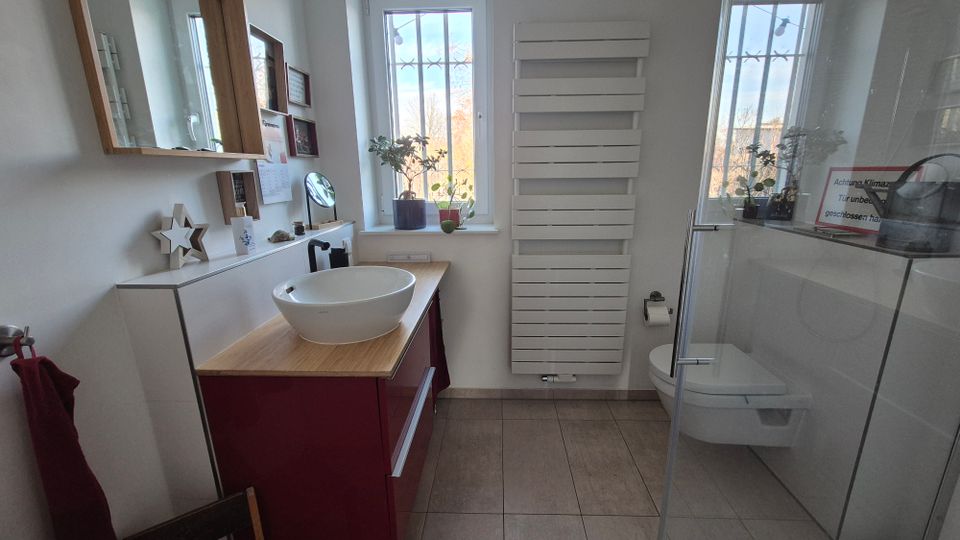 Modernes Badezimmer mit weißen Wänden und beigem Fliesenboden, ausgestattet mit einer roten Waschtischkommode mit ovalem Aufsatzwaschbecken, einem Spiegel mit Holzrahmen, einem Wandheizkörper und einer bodenebenen Glasduschkabine. Zwei Fenster mit Gitterstäben lassen Tageslicht herein, ergänzt durch Pflanzen auf den Fensterbänken.
