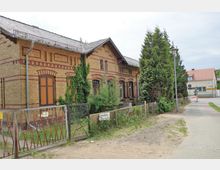 Backsteinbau mit gelb-roten Ziegelmustern, bedeckt von einem Satteldach, teils von Efeu bewachsen. Der Bau steht an einer gepflasterten Straße mit Vorgarten und umliegenden Wohnhäusern im Hintergrund.