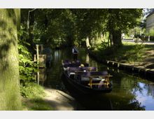 Eine Holz-Panoramabarkasse mit Tischen und lila Sitzpolstern fährt auf einem schmalen, von Bäumen gesäumten Kanal, gesteuert von einem stehenden Fährmann. Im Hintergrund und am Ufer sind grüne Vegetation und ein Weg sichtbar.