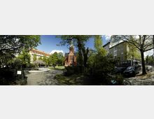 Ein kleiner Park mit einer gepflasterten Fläche, einem runden Springbrunnen und grünen Bäumen. Im Hintergrund steht eine rote Backsteinkirche mit gotischer Architektur, flankiert von mehrstöckigen Wohngebäuden.