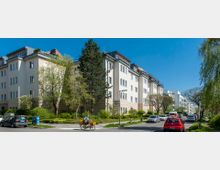 Wohnstraße mit mehrstöckigen, hellen Wohngebäuden in einer grünen Umgebung. Bäume und Büsche säumen die Straße, auf der parkende Autos sowie ein Postbote auf einem Lieferfahrrad zu sehen sind.