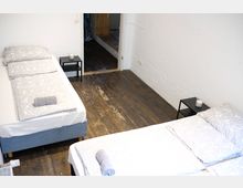 Ein einfach eingerichtetes Schlafzimmer mit zwei Einzelbetten, grauer Bettwäsche und dunklem Holzboden. Neben jedem Bett steht ein kleiner, schwarzer Nachttisch mit einer Kerze darauf, und im Hintergrund führt eine Tür zu einem angrenzenden Raum.