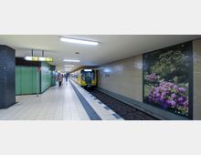 U-Bahn-Station mit gefliesten Wänden, einem gelben Zug, der am Gleis hält, und einem großen Wandbild mit Blumen und einem Gartenmotiv. Im Vordergrund ein heller gefliester Bahnsteig mit wenigen Passanten und Wegweisern an der Decke.