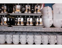 Regale mit gestapelten Metallkannen, großen weißen Tassen und kleineren, gestapelten Kaffeetassen mit blauem Logo, auf einer Arbeitsfläche aus Granit. Im Hintergrund ist ein blauer Wandbereich zu erkennen.