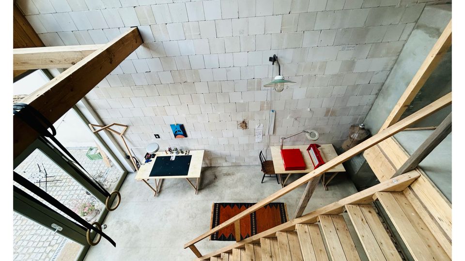 Innenansicht eines hohen, minimalistischen Raums mit hellen, kleinteiligen Wandfliesen, Sichtbetonboden und einer Holztreppe mit Geländer. Große bodentiefe Fenster lassen Tageslicht herein; darunter stehen ein schmaler Tisch, ein Stuhl und wenige weitere Möbel.