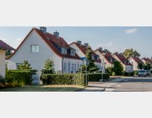 Eine ruhige Wohnsiedlung mit Reihenhäusern im Stil von Einfamilienhäusern, die weiße Fassaden und rote Satteldächer besitzen. Die Straße ist gesäumt von kleinen Vorgärten mit Hecken und Bäumen, daneben parken mehrere Autos.