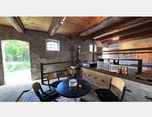 Rustikaler Innenraum eines umgebauten Scheunengebäudes mit groben Steinwänden, sichtbaren Holzbalken und Ziegelboden. Vor einem breiten Tresen mit Getränken stehen ein runder Tisch und mehrere Stühle; links öffnet sich ein großes Tor ins Grüne und kleine Fenster lassen Tageslicht herein.