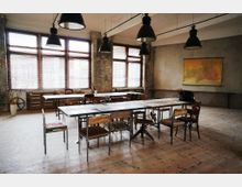 Ein geräumiger Raum mit industriellem Design, gekennzeichnet durch freiliegende Backsteinwände, große Fenster mit Holzjalousien und eine minimalistische Einrichtung. In der Mitte stehen lange Tische mit Holz- und Metallstühlen, darüber hängen schwarze Industrielampen, an der Wand eine große Landkarte.