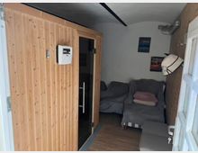 Ein kleiner Raum mit einer Holzsauna auf der linken Seite und einer Sitzecke auf der rechten Seite, die aus zwei mit grauen Decken abgedeckten Sesseln besteht. An der Wand befinden sich dekorative Poster, und ein geflochtener Teppich bedeckt den Boden.