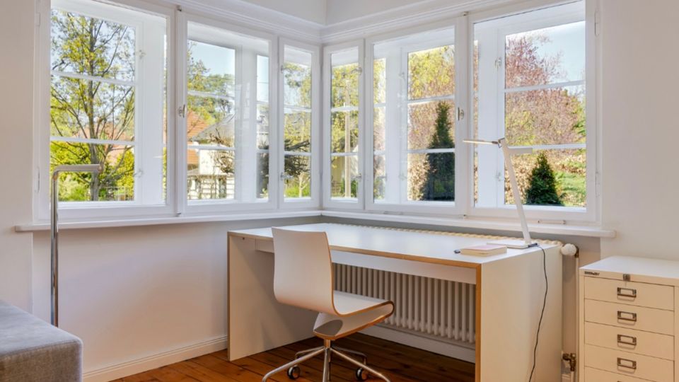 Heller Arbeitsbereich in einem Zimmer mit breiter Fensterfront über Eck und weißen Sprossenfenstern mit Blick ins Grüne. Unter den Fenstern steht ein schlichter weißer Schreibtisch mit Bürostuhl und Lampe, daneben ein schmaler Rollcontainer auf Holzdielenboden.