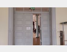 Große Doppeltür in einem historischen Gebäude, geöffnet mit Blick in einen Saal mit hohen Decken, einer verzierten Säule und Kronleuchtern. Über der Tür ist ein Notausgangsschild angebracht.