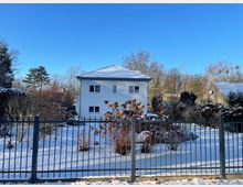 Blick auf ein freistehendes, zweigeschossiges Wohnhaus mit weißer Fassade und leicht verschneitem Walmdach in einem winterlichen Garten. Im Vordergrund verläuft ein dunkler Metallzaun, dahinter liegen schneebedeckte Beete und Sträucher sowie kahle Bäume unter blauem Himmel.