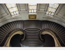 Große, geschwungene Doppeltreppe in einem historischen Gebäude mit stilisierten Metallgeländern, zentralem Podest und symmetrischer Gestaltung. Oberhalb befinden sich drei große Fenster mit Buntglas sowie eine verzierte goldene Wandplatte.