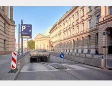 Einfahrt zu einer öffentlichen Tiefgarage mit einem blauen Parkhinweisschild im Vordergrund, flankiert von historischen Gebäuden mit aufwendiger Fassadenarchitektur und einer Kuppel im Hintergrund. Die Szene zeigt eine städtische Umgebung bei Tageslicht.