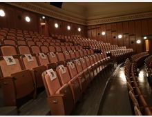 Reihenweise geordnete, gepolsterte Sitzplätze in einem historischen Zuschauerraum mit Holzwänden und rundlichen Wandlampen, die weiches Licht spenden; auf einigen Sitzplätzen sind bedruckte Zettel angebracht.